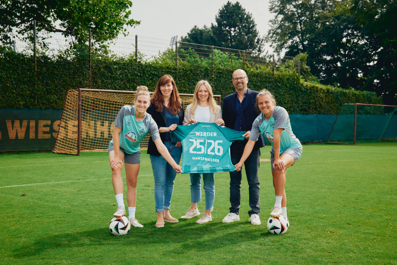 Zwei Spielerinnen von Werder Bremen und drei Mitarbeitende von hanseWasser halten gemeinsam ein Werdertrikot mit hanseWasser Schriftzug.