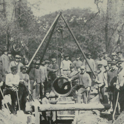 Schwarzweiß-Foto von 1893, viele Menschen mit Schaufeln stehen um eine Baustelle